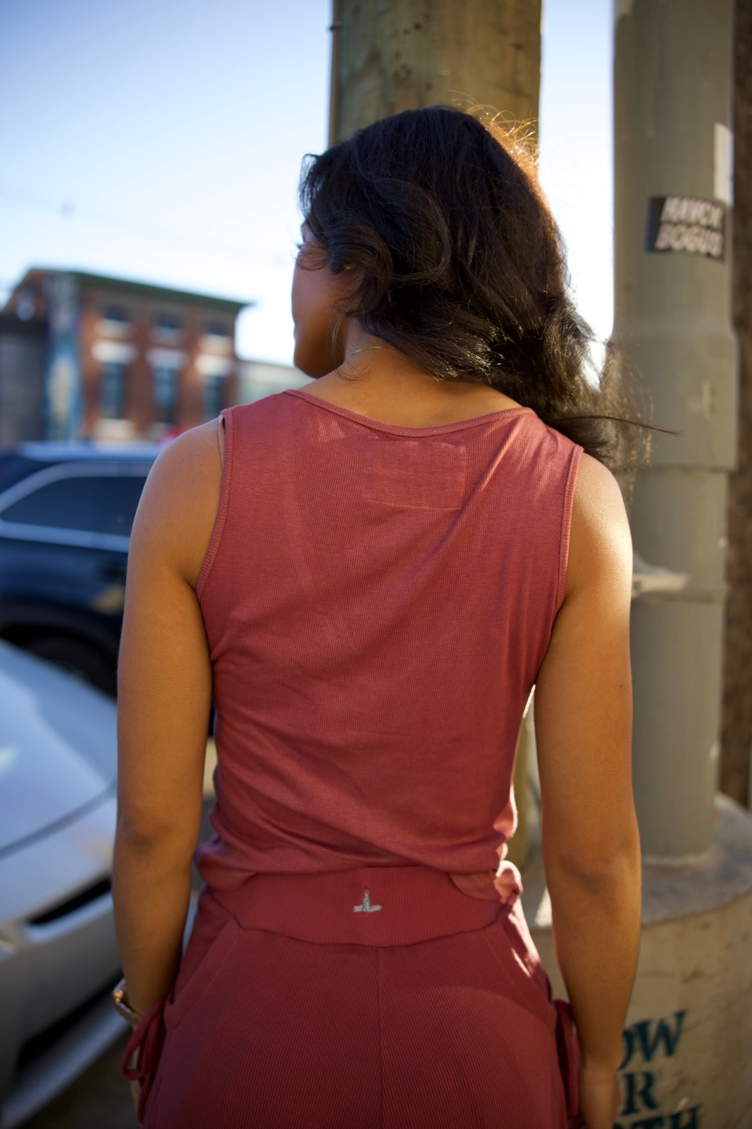 Person wearing a pink sleeveless top and pants standing outdoors with buildings and a car in the background.
