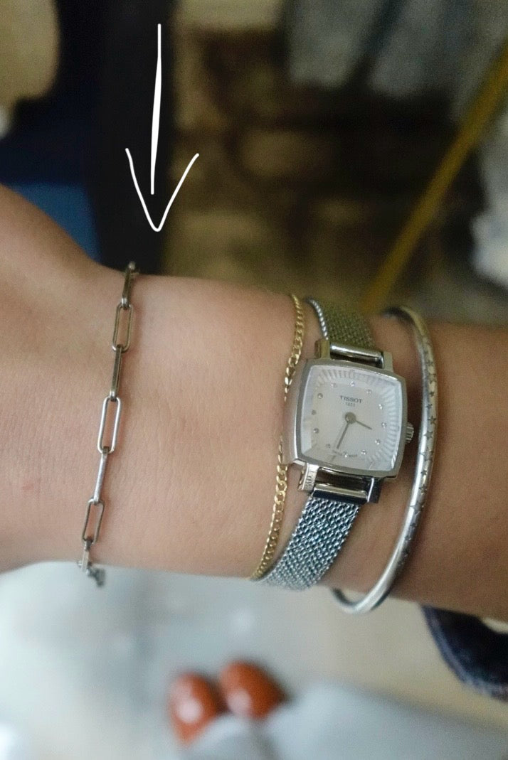 Wrist with multiple bracelets and a watch, with an arrow pointing to the watch.