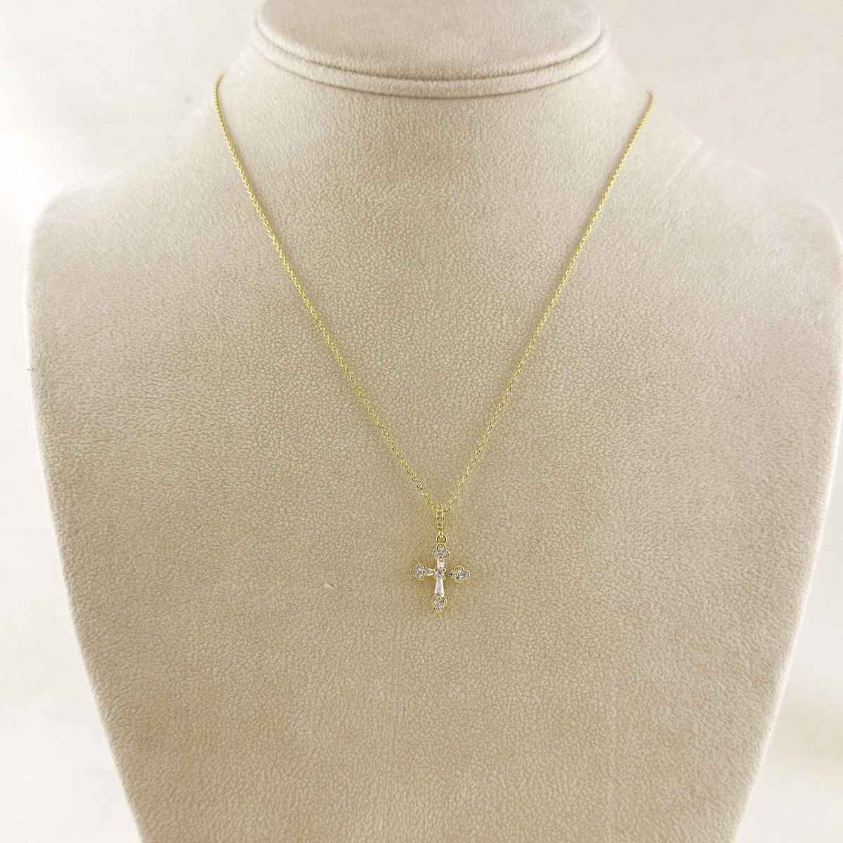 A delicate gold chain necklace with a small cross pendant, displayed on a cream-colored jewelry bust.