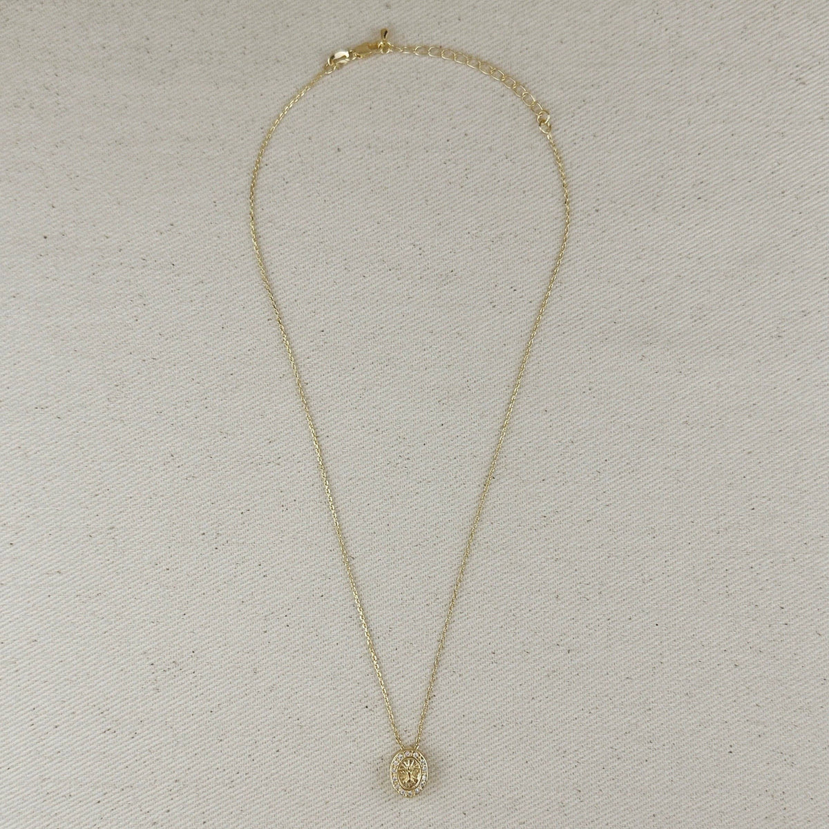 A delicate gold chain necklace with a small oval pendant, laid flat on a textured beige fabric background.