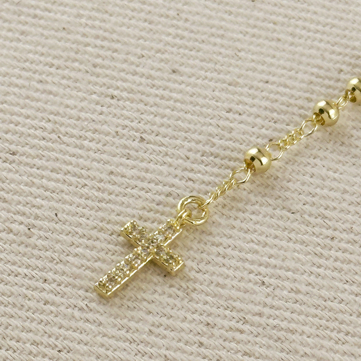 A gold cross pendant with sparkling clear stones, attached to a gold beaded chain, displayed on a textured beige fabric background.