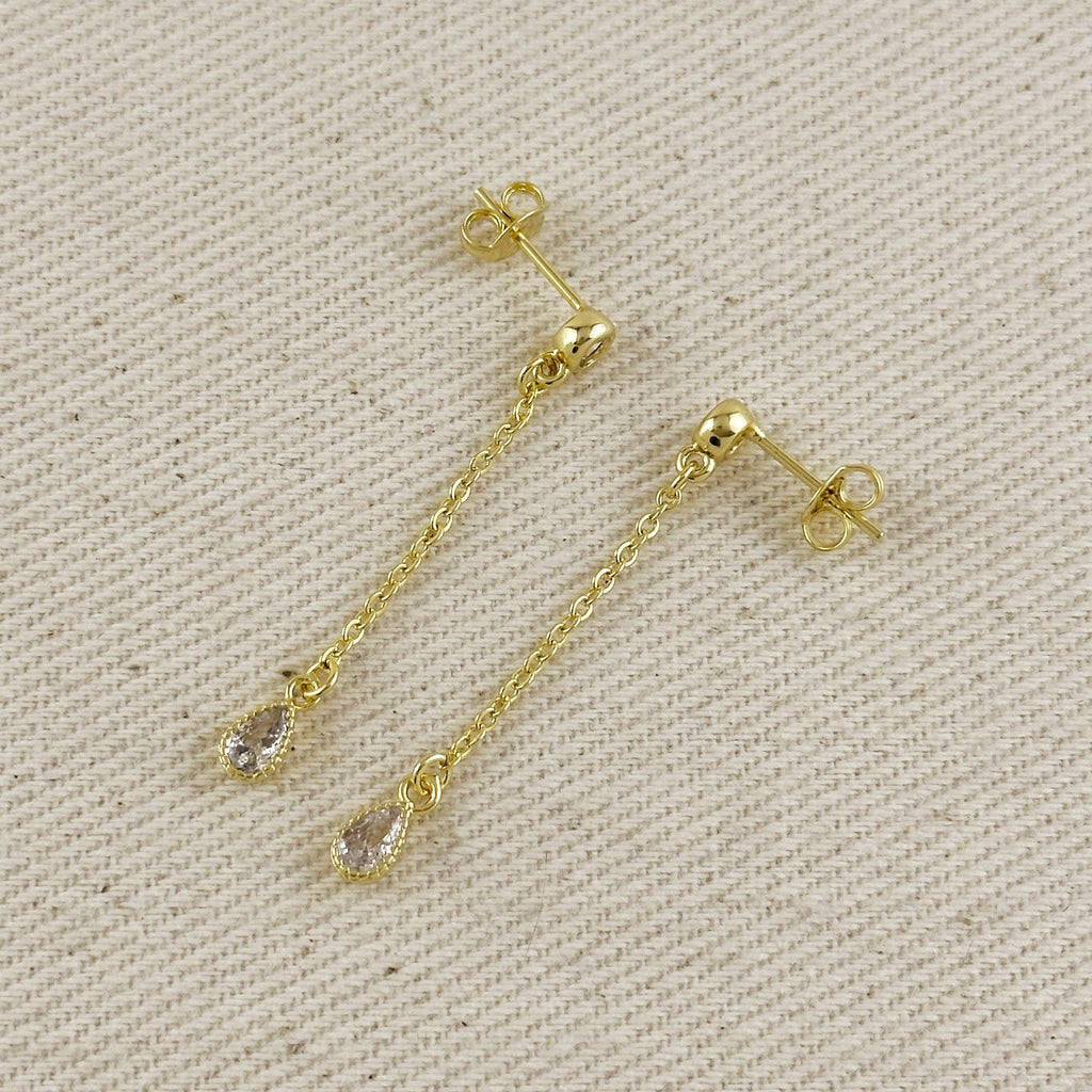 A pair of gold dangle earrings with teardrop-shaped clear gemstones, displayed on a textured beige fabric background.