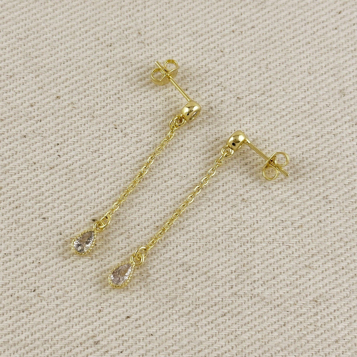 A pair of gold dangle earrings with teardrop-shaped clear gemstones, displayed on a textured beige fabric background.