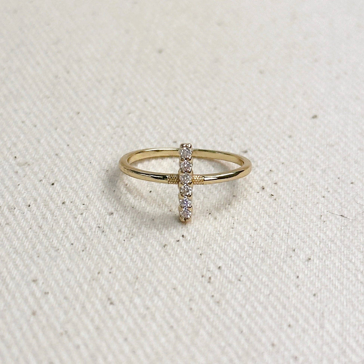 A delicate gold ring featuring a vertical bar studded with small clear stones, resting on a textured off-white fabric background.