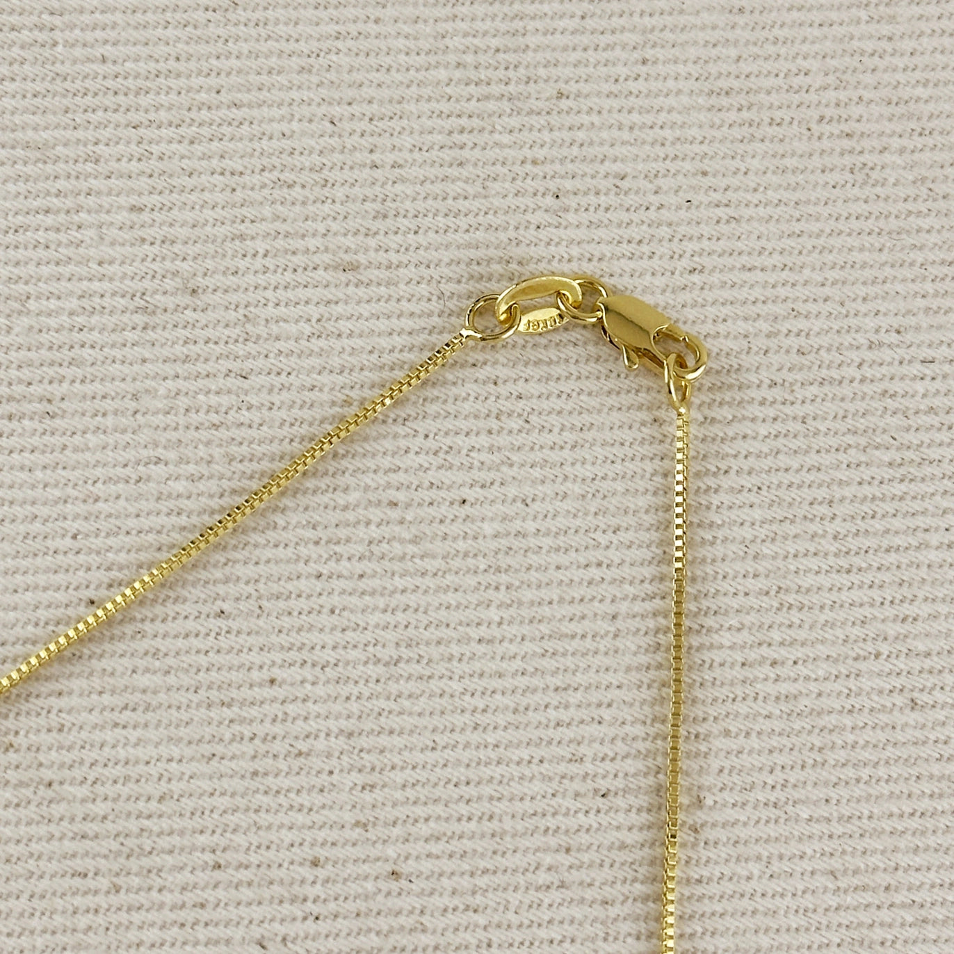 Close-up of a gold chain necklace clasp and extender on a textured beige fabric background.