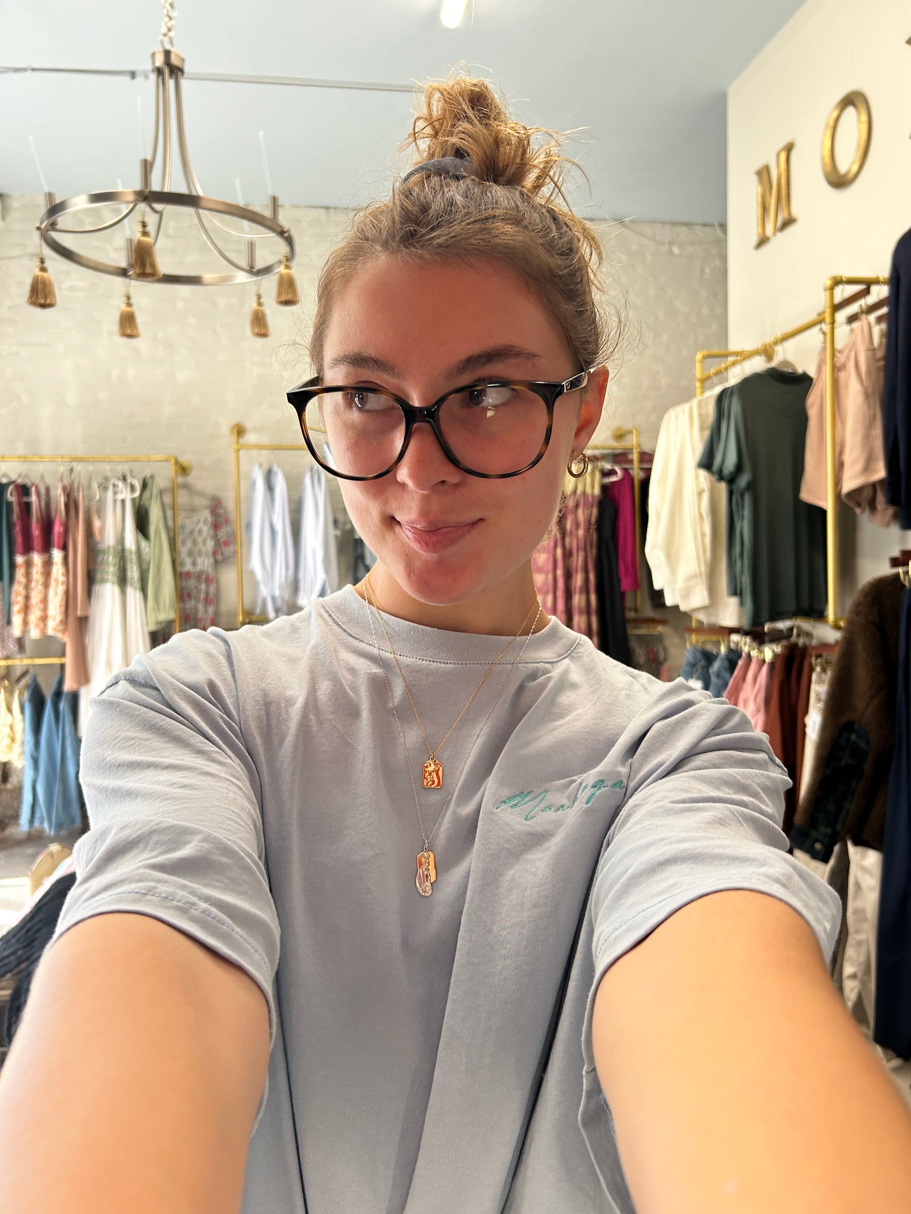 Person wearing glasses and a light gray shirt in an indoor setting with reflective surfaces.