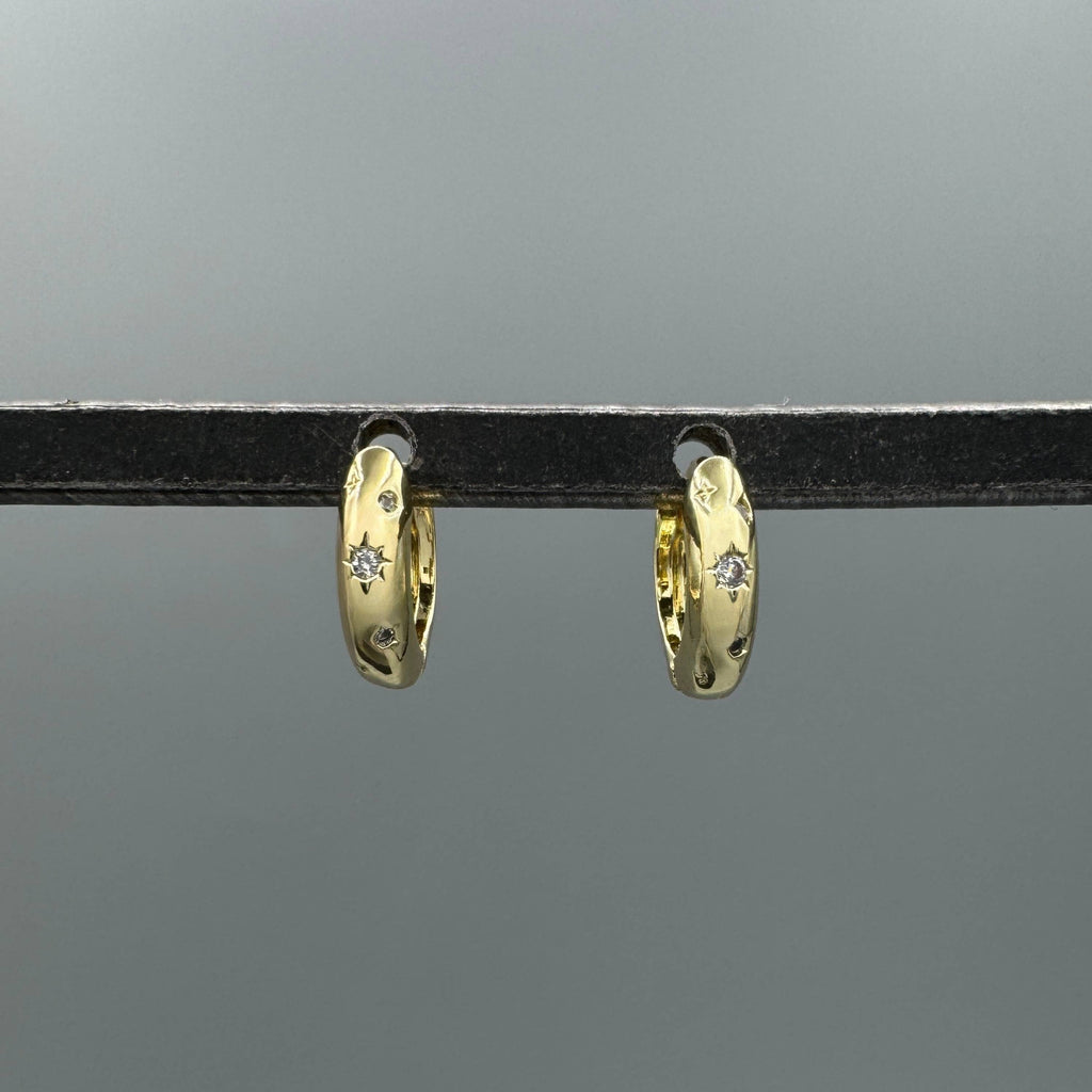 A pair of small gold huggie hoop earrings with tiny clear stones, hanging from a black display bar against a gray background.