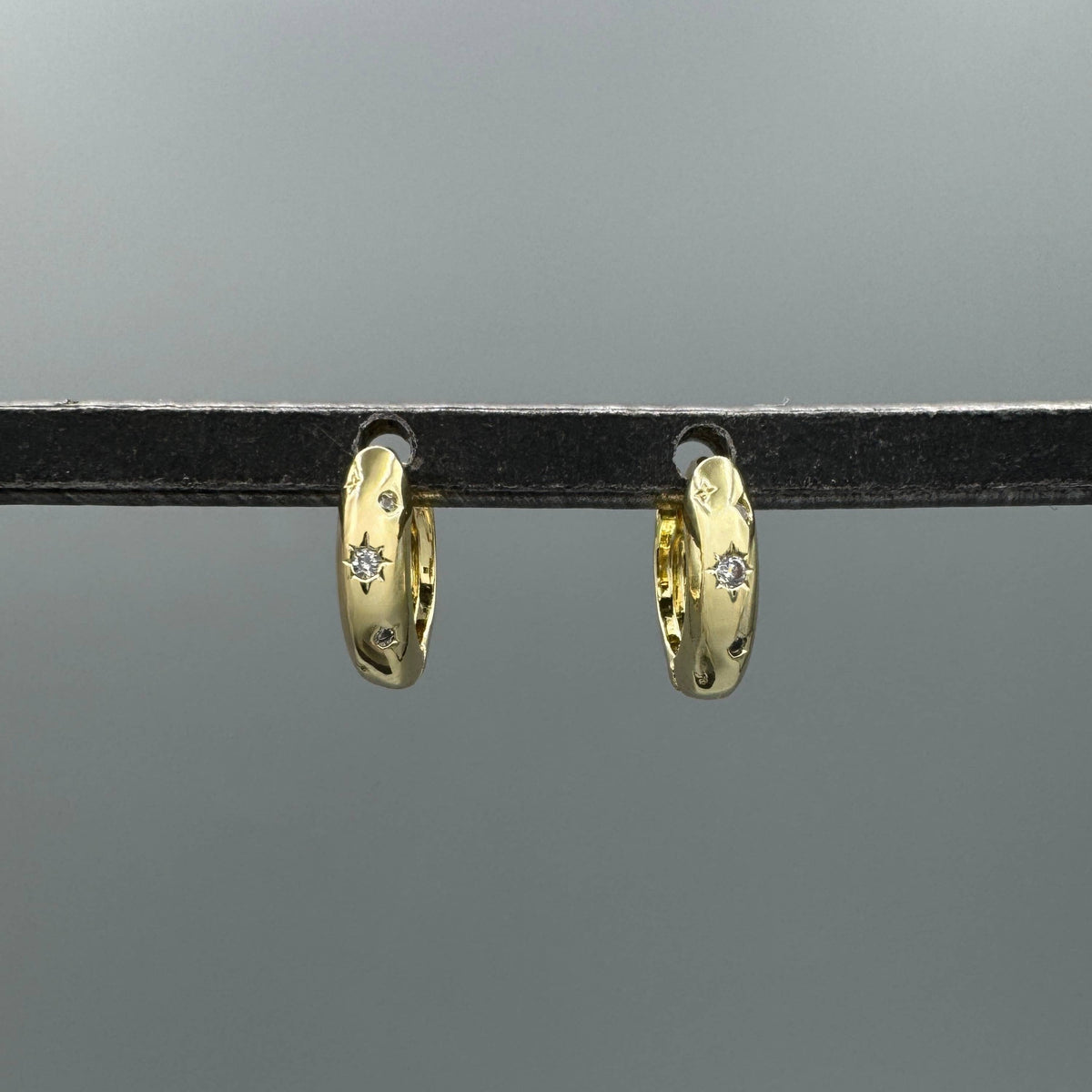 A pair of small gold huggie hoop earrings with tiny clear stones, hanging from a black display bar against a gray background.
