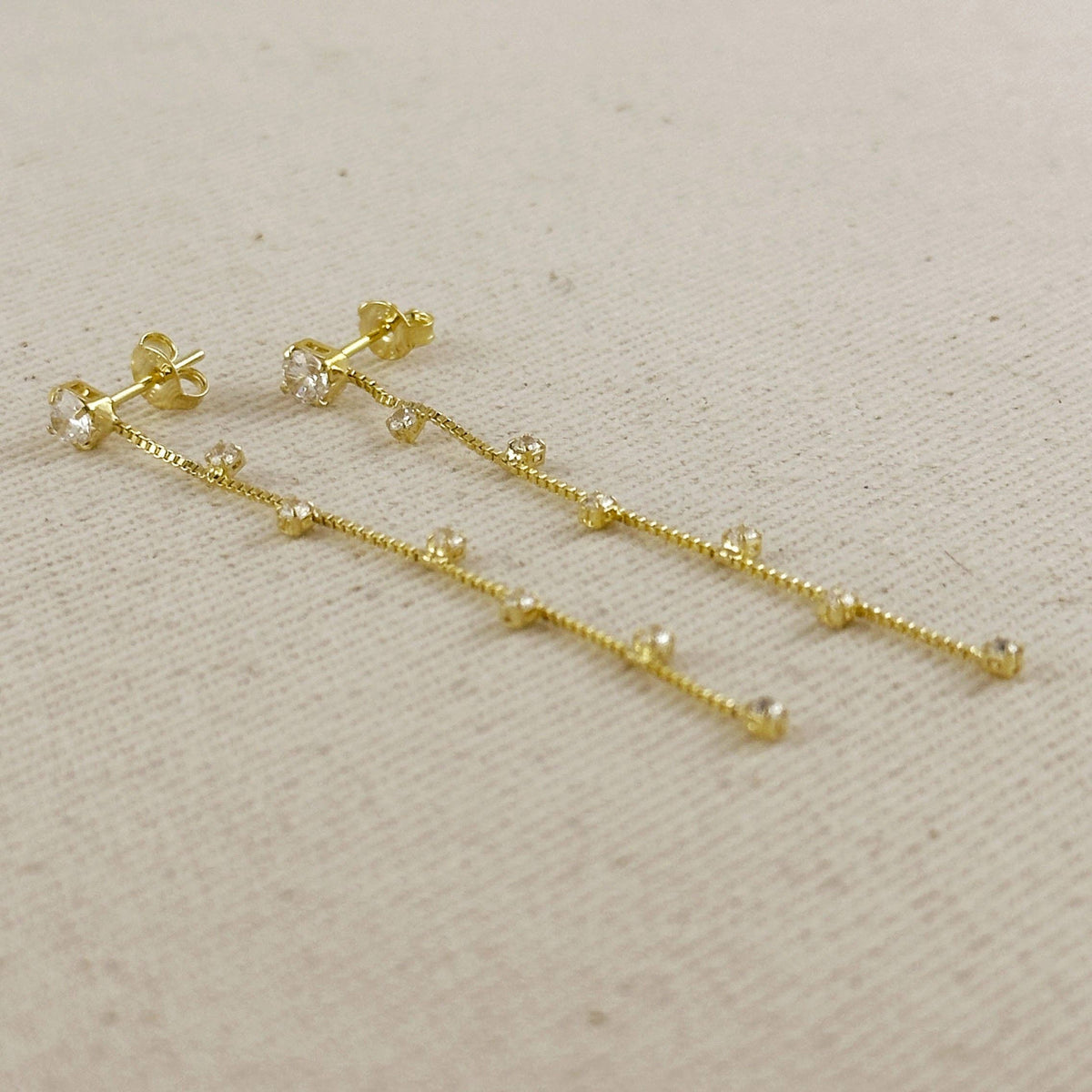 A pair of gold dangle earrings with small clear gemstones set along a delicate chain, displayed on a textured beige fabric background.