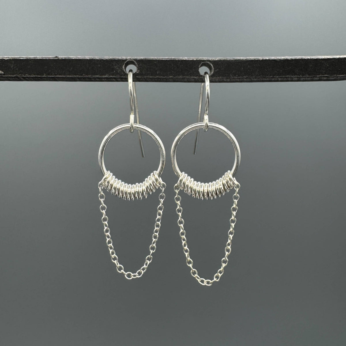 A pair of silver dangle earrings with circular hoops, textured wire wrapping at the top, and delicate chain drops, hanging from a black display bar against a dark gray background.