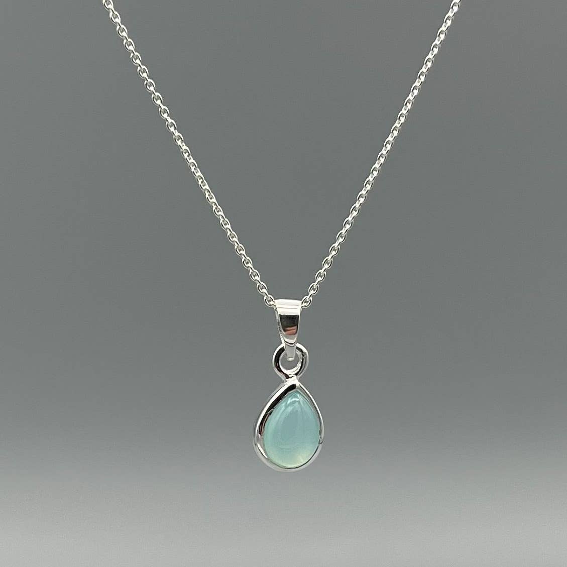 A silver chain necklace with a teardrop-shaped pendant featuring a milky, pale greenish-blue stone in a polished silver setting, shown against a neutral gray background.
