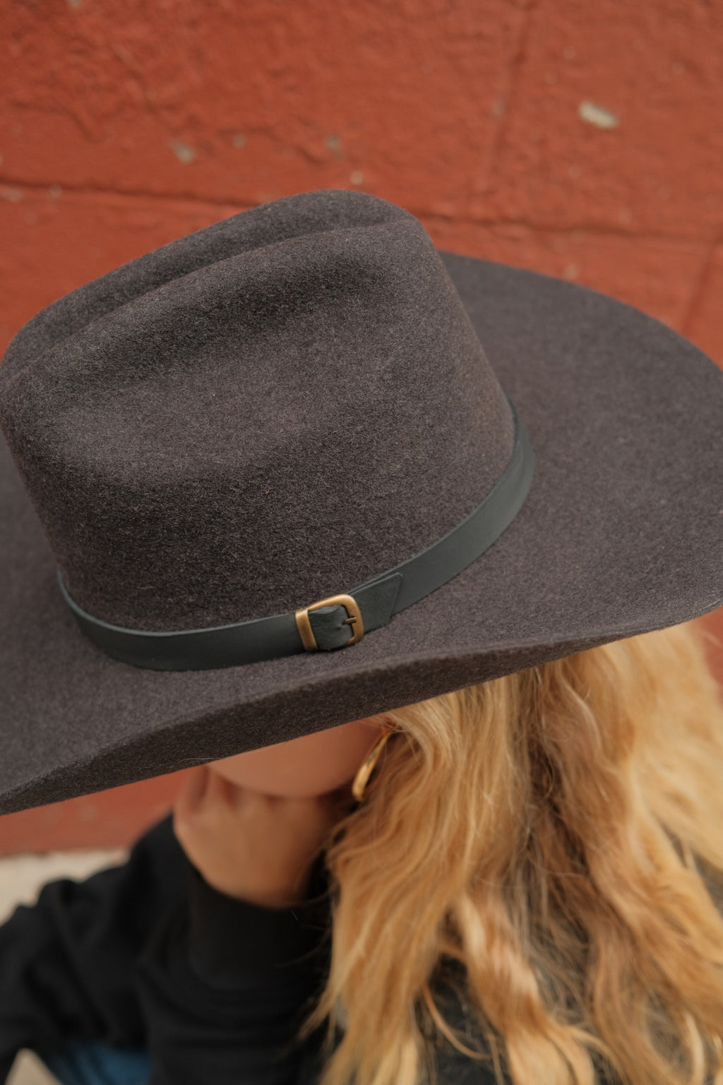 A close-up of a person wearing a dark brown felt cowboy hat with a black leather band and gold buckle, set against a textured red brick wall.