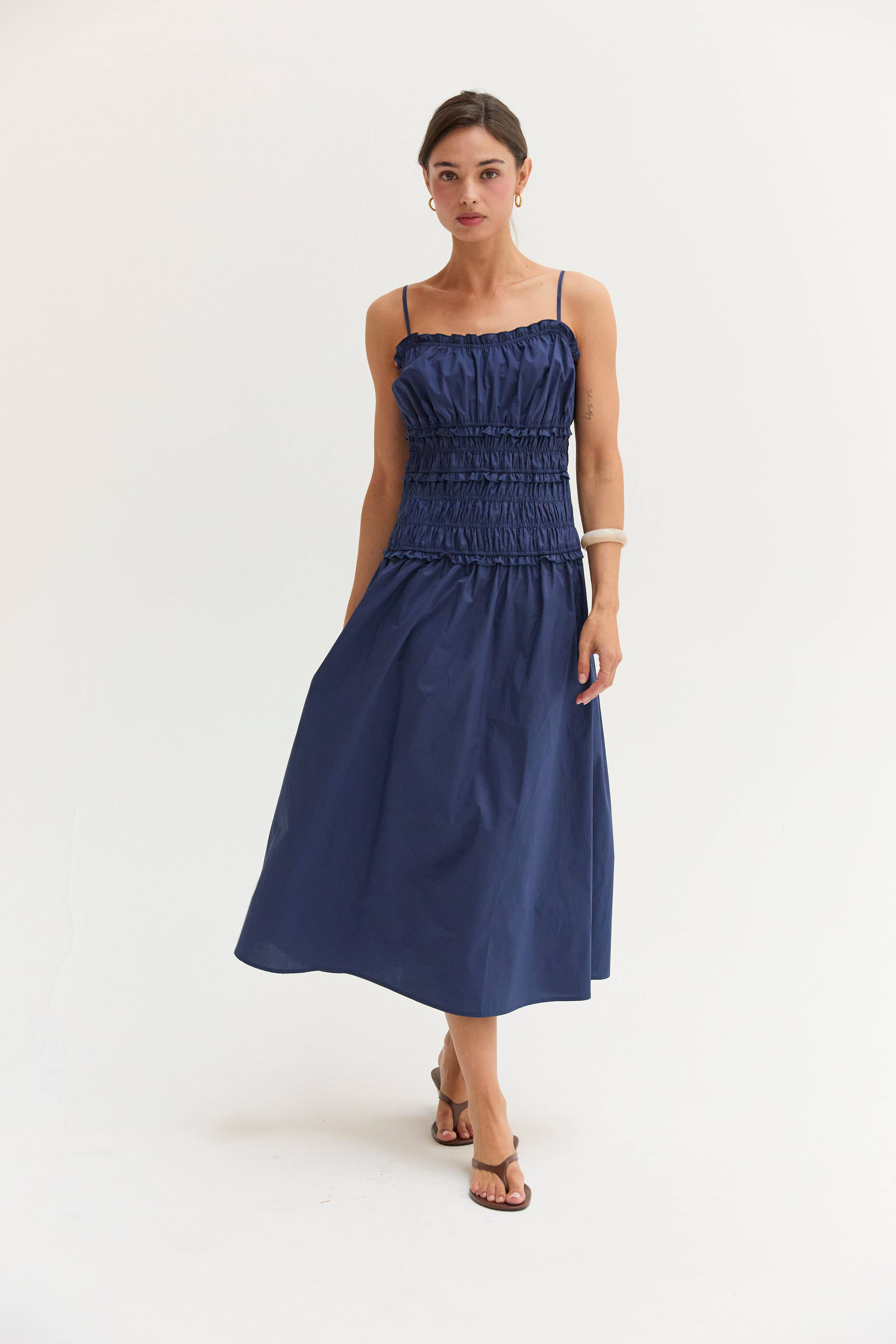 A woman modeling a navy blue midi dress with thin straps, a smocked bodice, and a flared skirt against a plain white studio background.