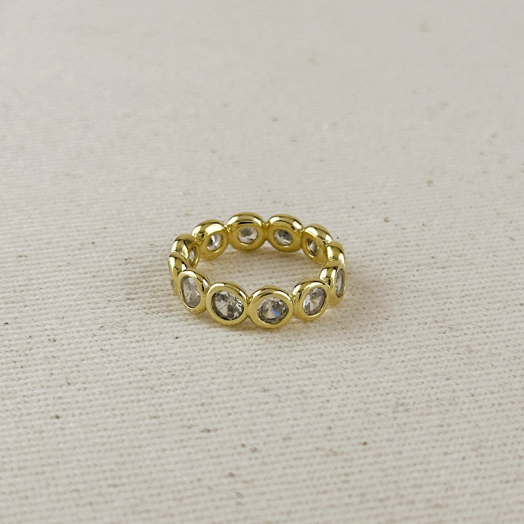 A gold eternity-style ring featuring multiple round clear gemstones set in a polished band, photographed on a textured beige fabric background.
