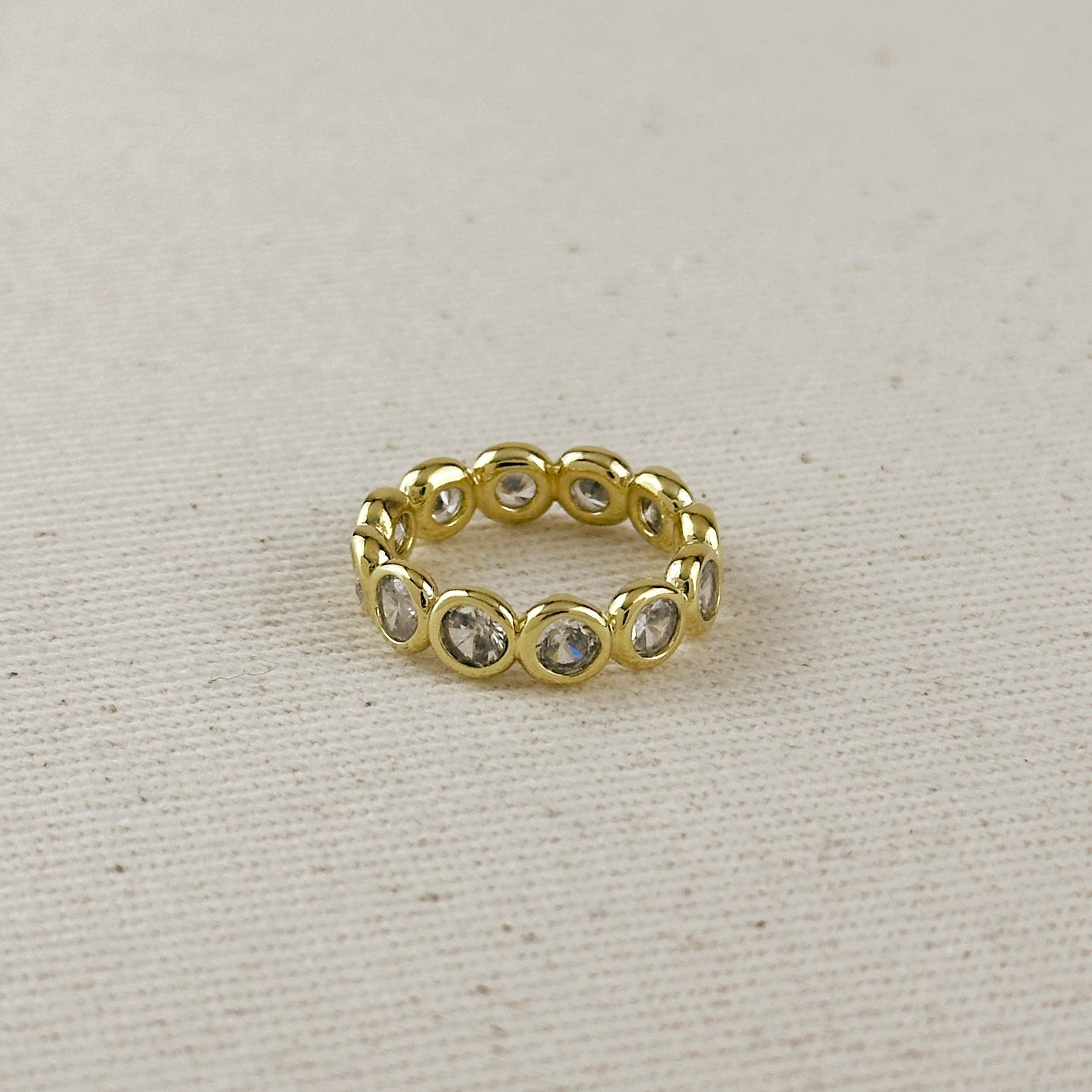 A gold eternity-style ring featuring multiple round clear gemstones set in a polished band, photographed on a textured beige fabric background.