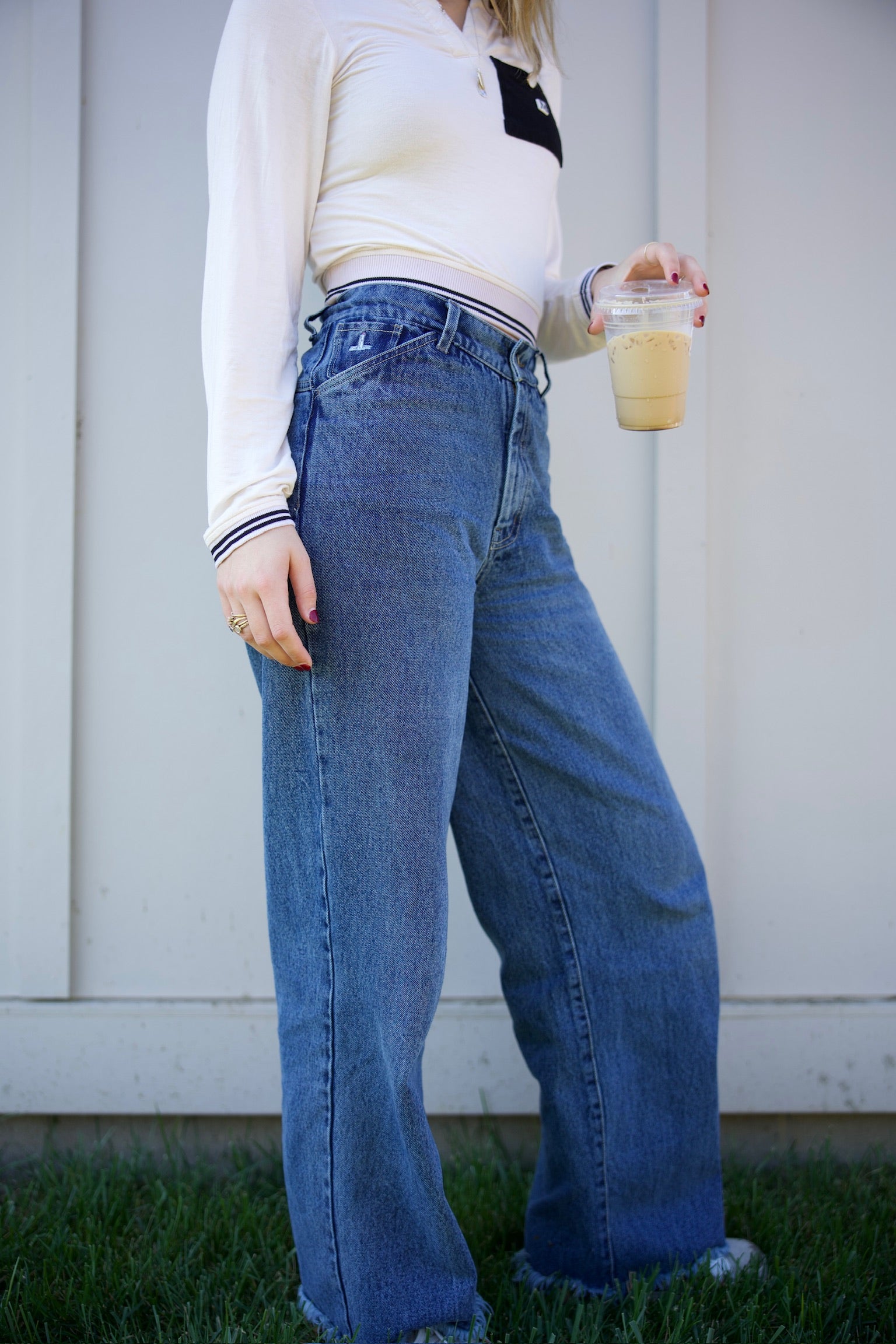 Person wearing blue jeans and a white sweater holding a drink outdoors.