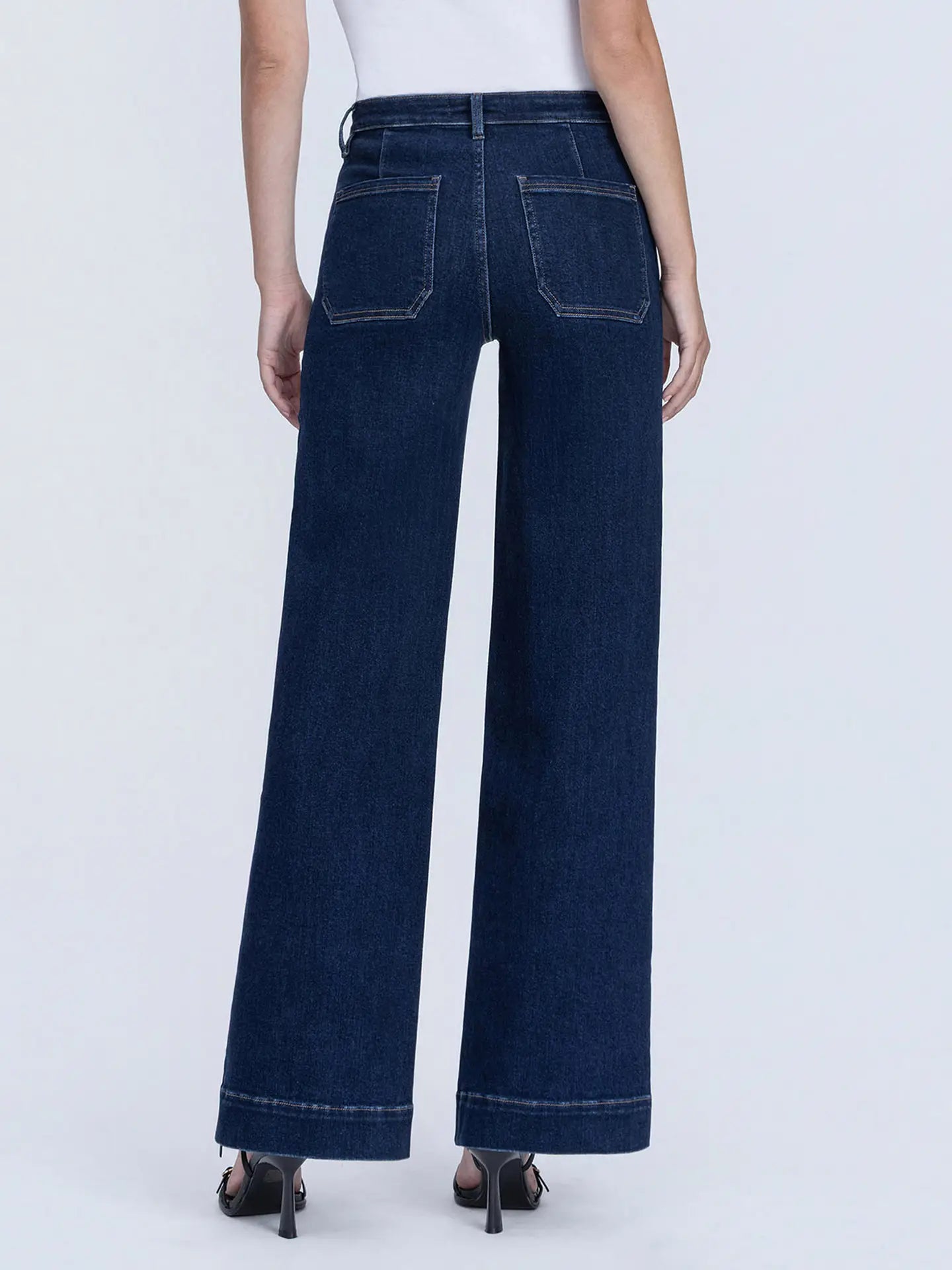 A person shown from the back wearing dark blue wide-leg jeans with contrast stitching, paired with a white top and black heels, against a light gray studio background.