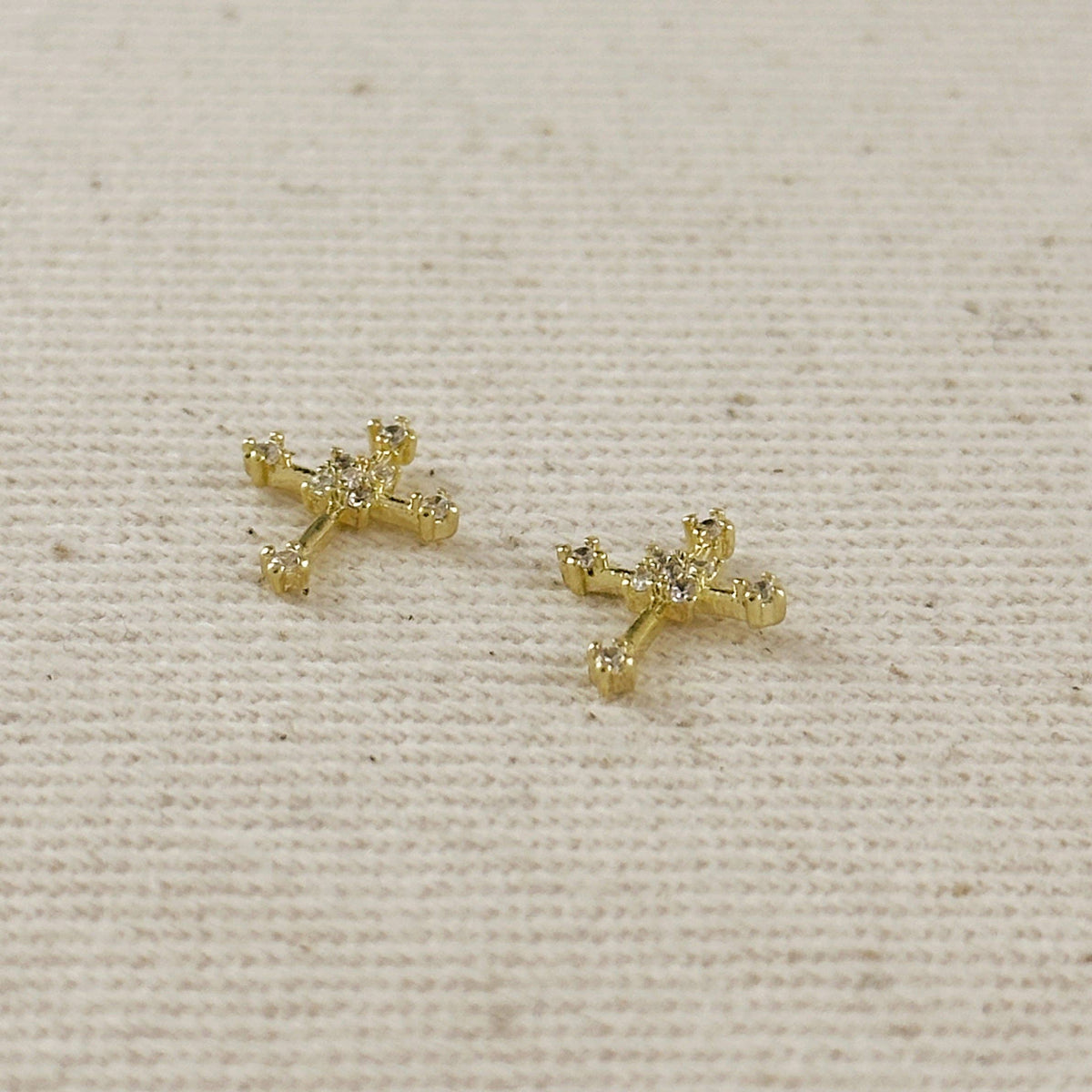 Gold cross-shaped earrings with small diamonds on a beige fabric background