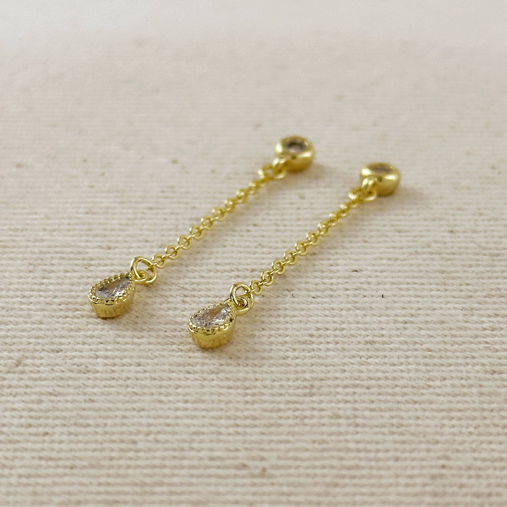 A pair of gold dangle earrings with teardrop-shaped clear gemstones, displayed on a textured beige fabric background.