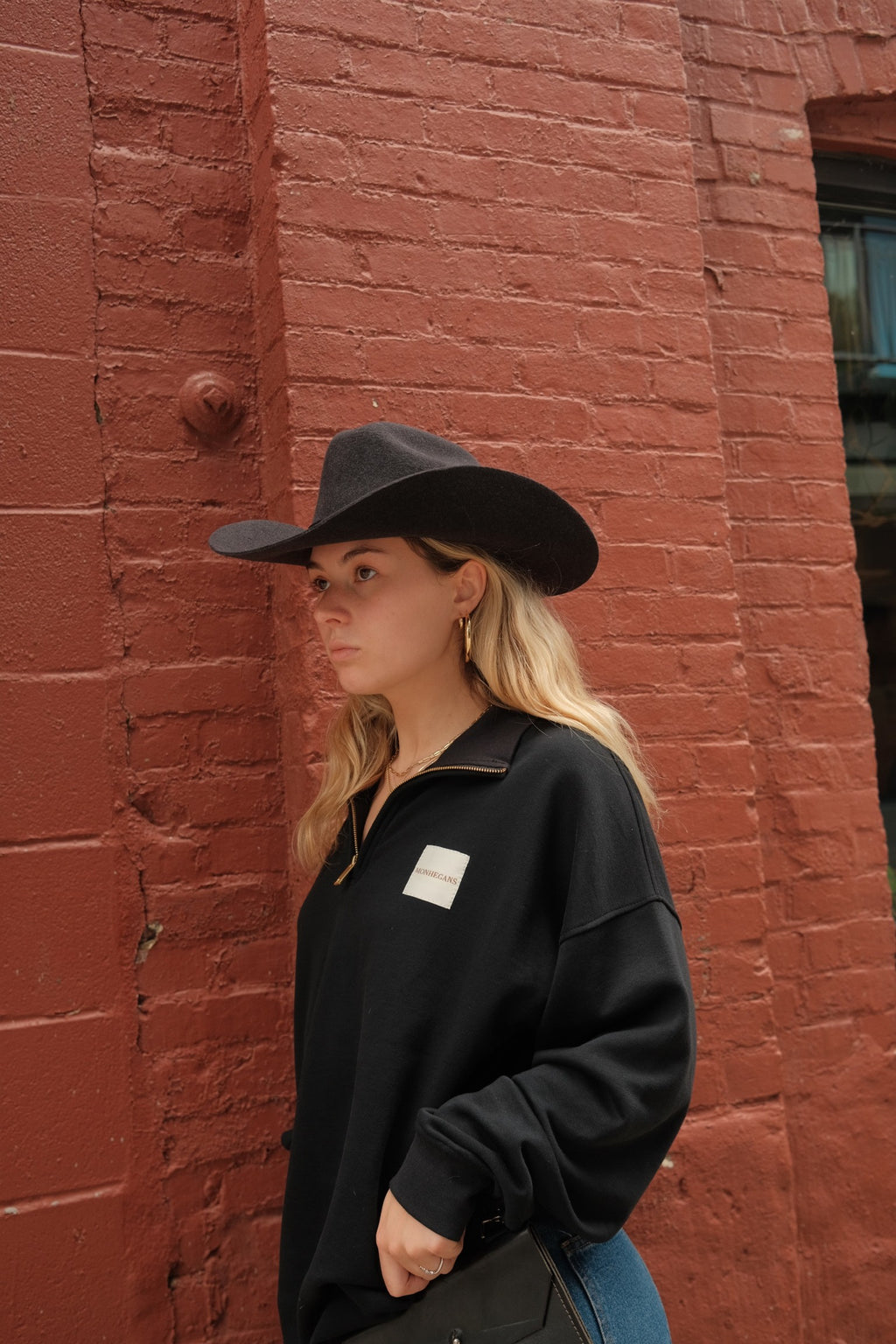 A woman wearing a black wide-brim hat and a black half-zip sweatshirt with a small white square patch on the chest, standing against a red brick wall.