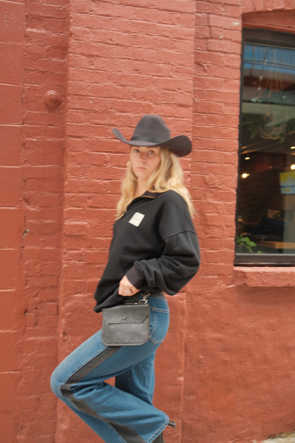A woman posing in front of a red brick wall, wearing a black cowboy hat, a black sweatshirt, blue jeans with black contrast panels, and carrying a small black shoulder bag.