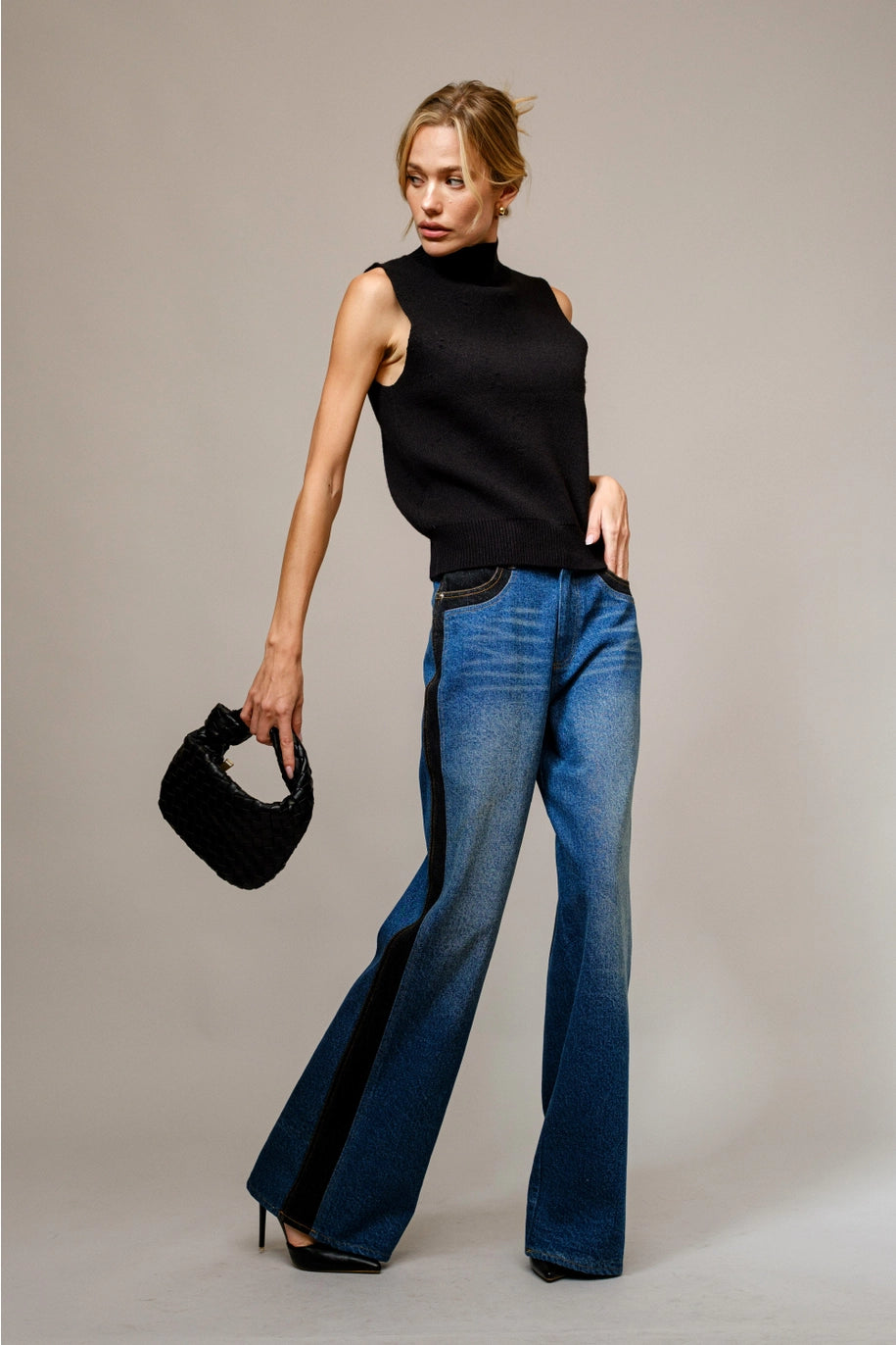 Woman wearing a black sleeveless top and blue jeans holding a black handbag against a neutral background