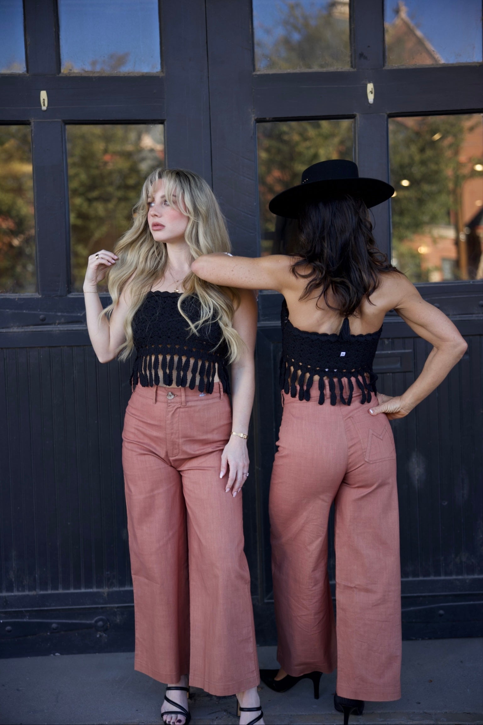 Two women wearing matching outfits with black tops and pink pants, standing in front of a dark door.